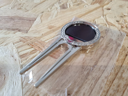 A silver metal golf divot tool with a dark oval center and a rhinestone-studded circular head, resting on a clear plastic bag over a light wood particle board surface.
