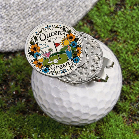 A golf flag holder with a round enamel pin design reading 'Queen of the Green' and featuring golf-themed illustrations, shown attached to a silver metal flag clip on a white golf ball over grass.
