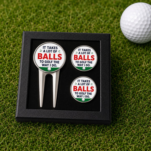 A golf accessory gift set in a black presentation box, featuring a silver metal golf divot tool and two matching round metal magnets or pins with the text 'IT TAKES A LOT OF BALLS TO GOLF THE WAY I DO.' The set is displayed on a green grass surface with a white golf ball in the background.